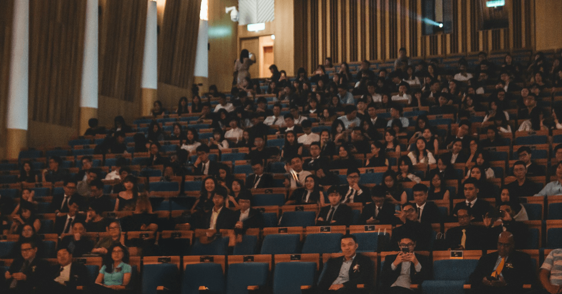 Audience seated in a tiered auditorium with blue chairs and wooden wall panels.